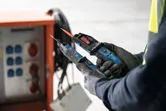 Persona, kas valkā drošības aprīkojumu, izmanto sprieguma testeri elektrības paneļa tuvumā.