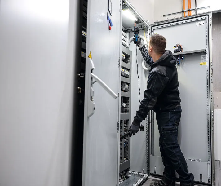 Persona, kas valkā drošības aprīkojumu, pārbauda elektrisko paneli ar sprieguma testeri.