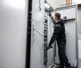 Persona, kas valkā drošības aprīkojumu, pārbauda elektrisko paneli ar sprieguma testeri.