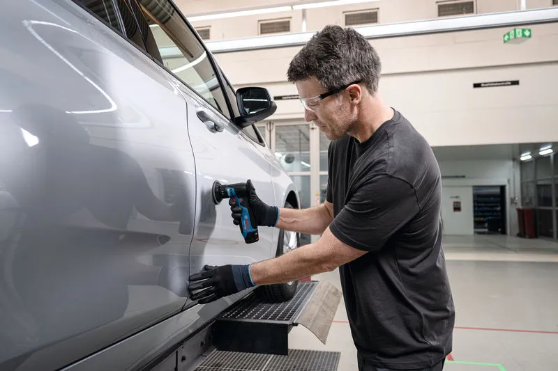 Persona, kas valkā drošības aprīkojumu, slīpē automašīnas durvis, izmantojot ekscentrisko slīpmašīnu.