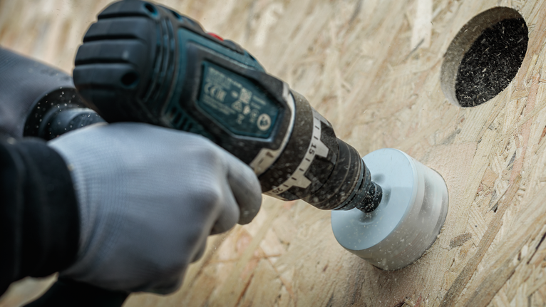 Cilvēks drošības aprīkojumā ar caurumzāģi urbj saplāksnī apaļu caurumu.