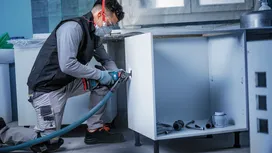 شخص يرتدي معدات السلامة يستخدم أداة كهربائية لقطع فتحة في خزانة المطبخ.