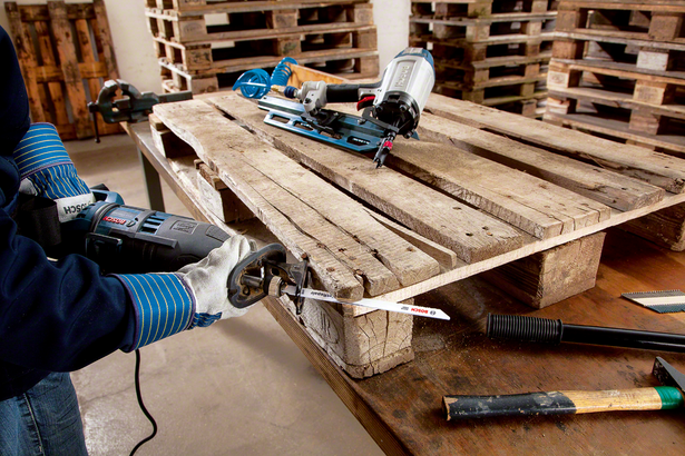 Worker wearing safety equipment uses a reciprocating saw to cut through a wooden pallet.