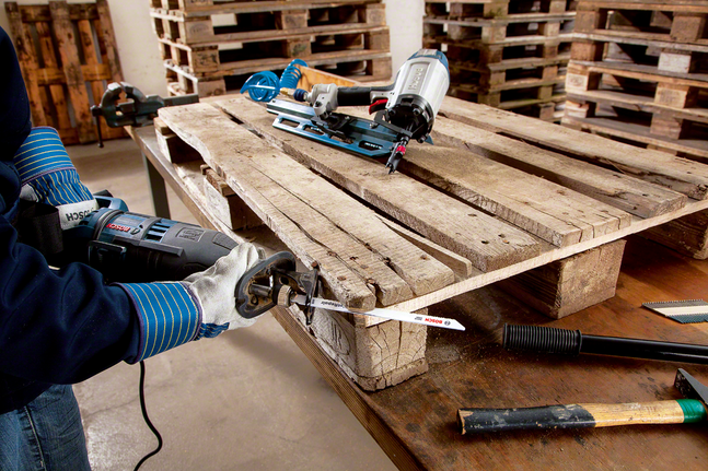 Worker wearing safety equipment uses a reciprocating saw to cut through a wooden pallet.