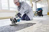 A person wearing safety equipment grinds a concrete floor with a power tool.
