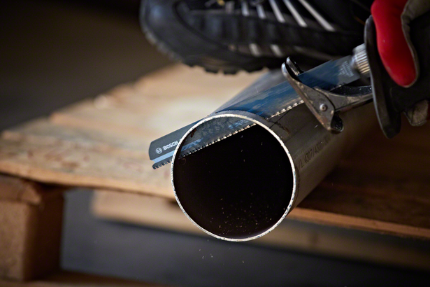 Worker wearing safety equipment uses reciprocating saw to cut a metal pipe.