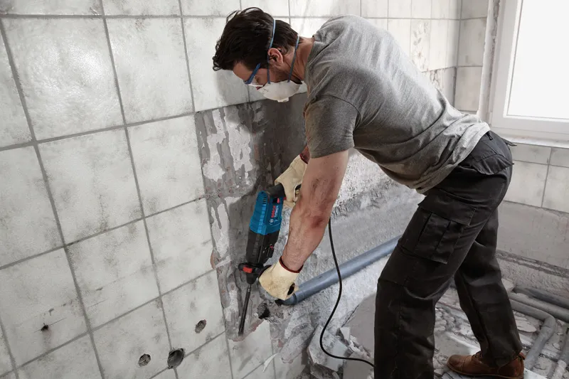 A person wearing safety equipment uses a power tool to remove tiles from a wall.