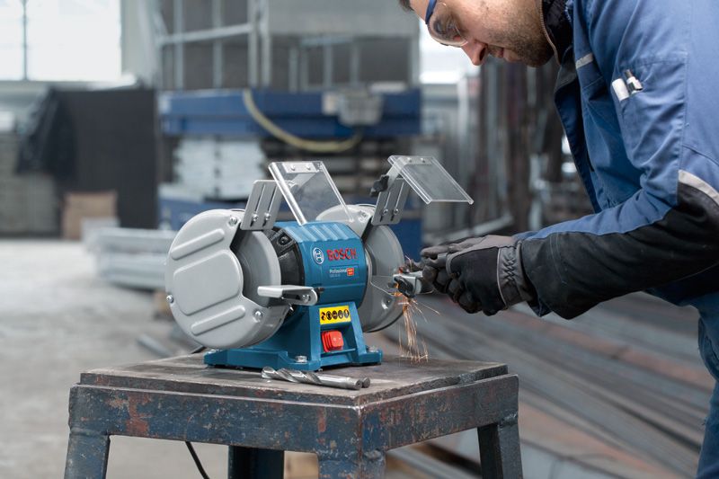 A person wearing safety equipment grinds metal on a double-wheeled bench grinder.