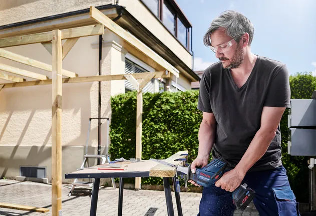 Person wearing safety equipment cuts wood with a cordless saw outdoors.