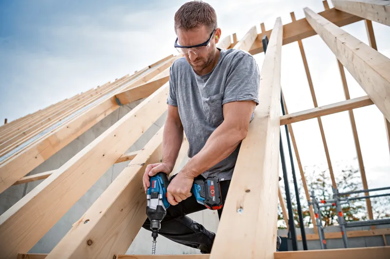 A person wearing safety equipment fastens a screw into a wooden frame using an impact wrench.