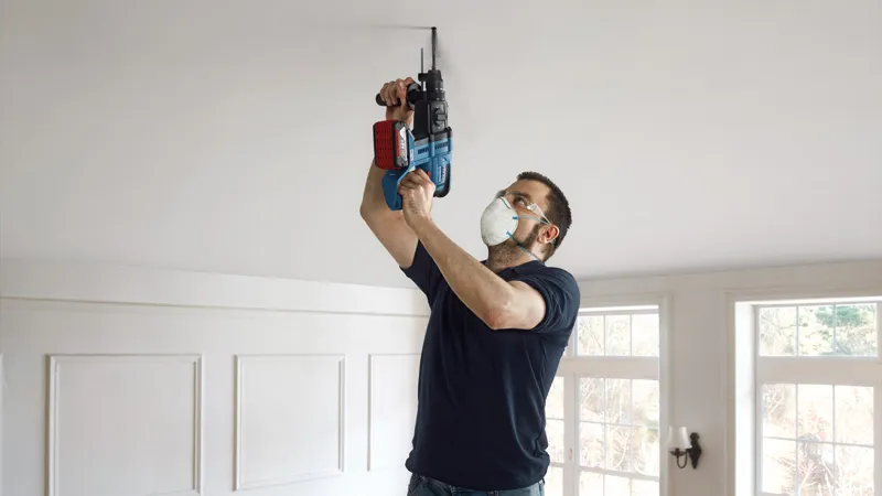 A person wearing safety equipment drills into a ceiling with a power tool.