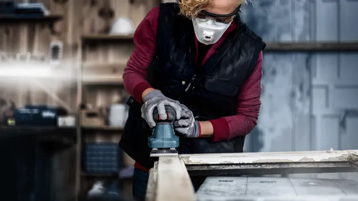 Person wearing safety equipment sands wood with a power tool in a workshop.