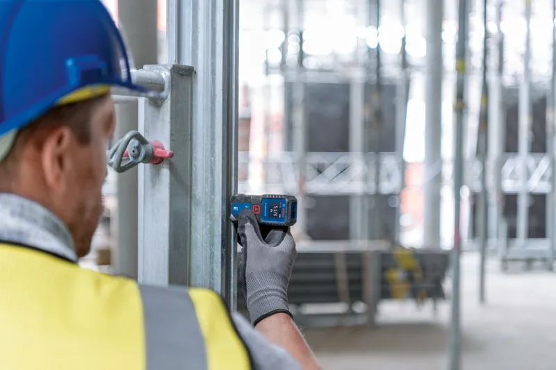 A person wearing safety equipment measures a metal beam with a laser measure.