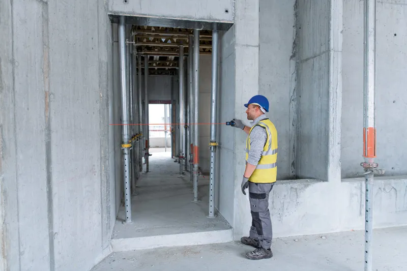 A person wearing safety equipment uses a laser measure to check distance in a concrete structure.