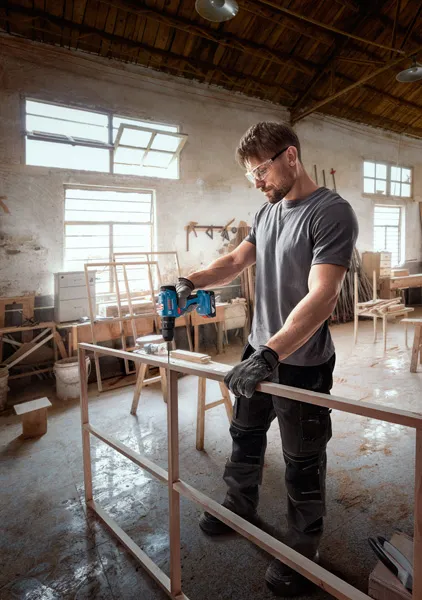 A person wearing safety equipment drills into a wooden frame in a workshop.