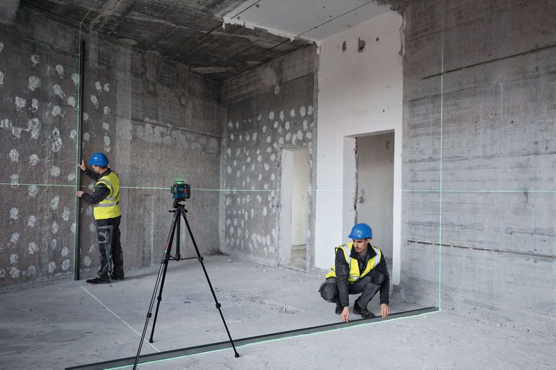 Two people wearing safety equipment align walls using a laser leveling tool in a building under construction.