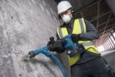 A person wearing safety equipment uses a cordless demolition hammer on a concrete wall.
