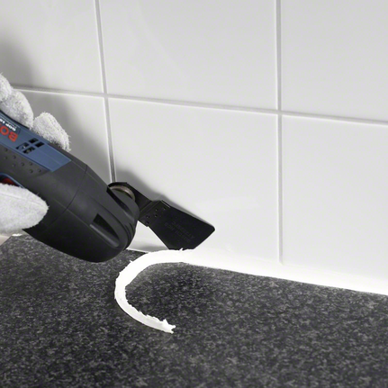 Gloved hand uses a multitool to remove silicone sealant from a tiled surface.