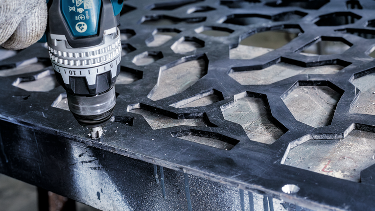 Person wearing safety equipment uses a drill to make a hole in a patterned metal sheet.