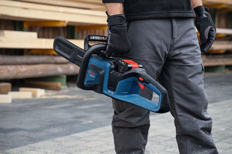 A person wearing safety equipment holds a cordless chainsaw at a lumber yard.