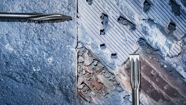 Two chisel drill bits removing concrete from a wall surface.