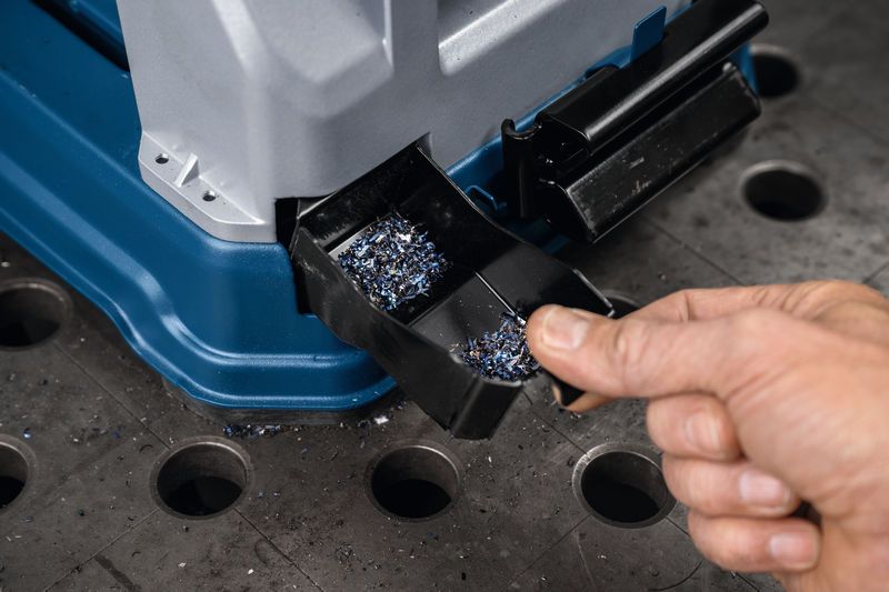 A person empties a metal chip tray from a metal cut-off saw on a workbench.