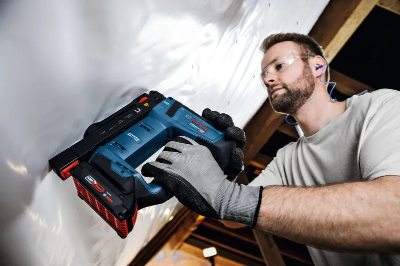 Person wearing safety equipment uses a cordless nail gun to fasten material to a ceiling.