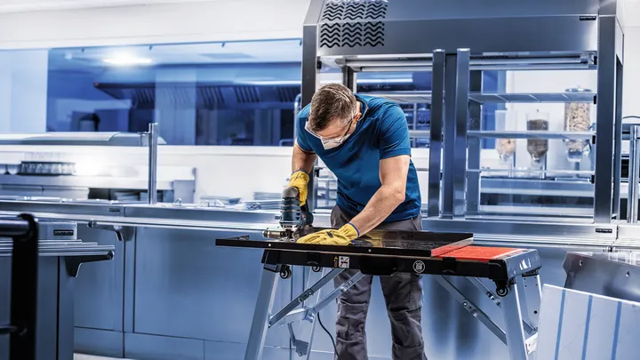Person wearing safety equipment uses a power tool to cut metal in a commercial kitchen.