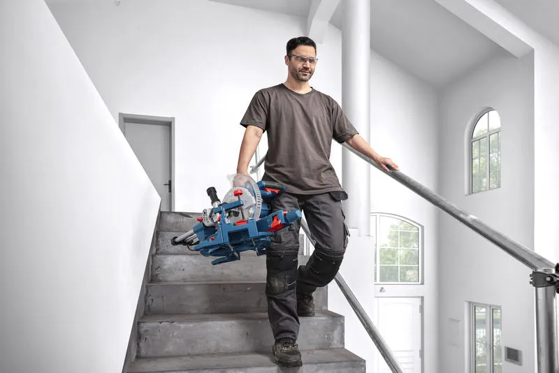 A person wearing safety equipment carries a mitre saw while walking down stairs.