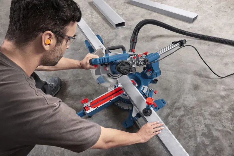 A person wearing safety equipment uses a mitre saw to cut a long metal beam.