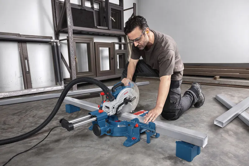 A person wearing safety equipment uses a mitre saw to cut a metal beam in a workshop.