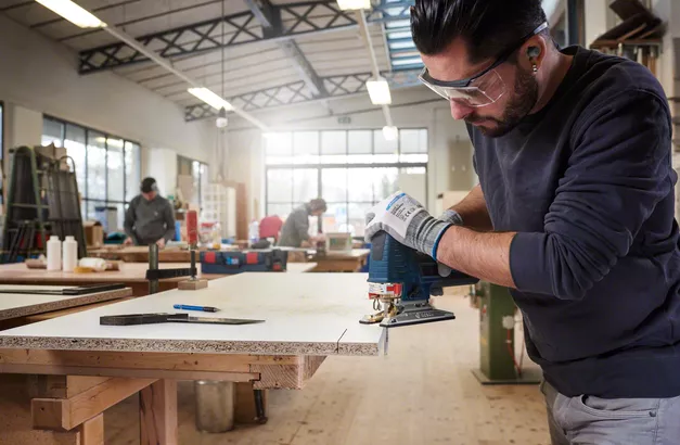 Une personne portant un équipement de sécurité utilise une scie sauteuse pour couper des panneaux de particules dans un atelier.