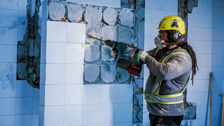 Une personne portant un équipement de sécurité retire des carreaux d'un mur à l'aide d'un marteau de démolition.