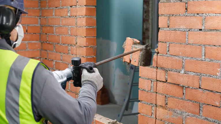 Une personne portant un équipement de sécurité retire des briques d’un mur à l’aide d’un outil de démolition.