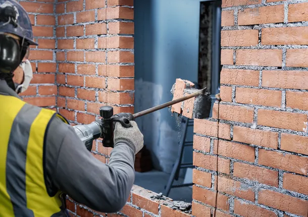 Une personne portant un équipement de sécurité brise un mur de briques à l'aide d'un outil de démolition.