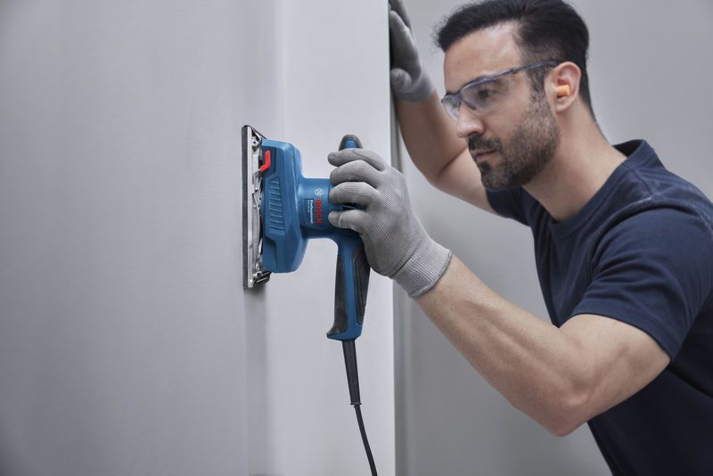 Une personne portant un équipement de sécurité ponce un mur avec une ponceuse orbitale.