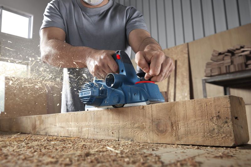 Une personne portant un équipement de sécurité rabote une poutre en bois, des copeaux de bois volent.