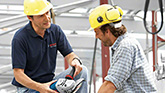Two men in hard hats are discussing at a construction site. One is pointing at a shoe.