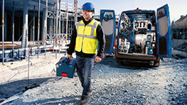 Un ouvrier du bâtiment en gilet de sécurité jaune et casque bleu porte une boîte à outils et se dirige vers une camionnette bleue.