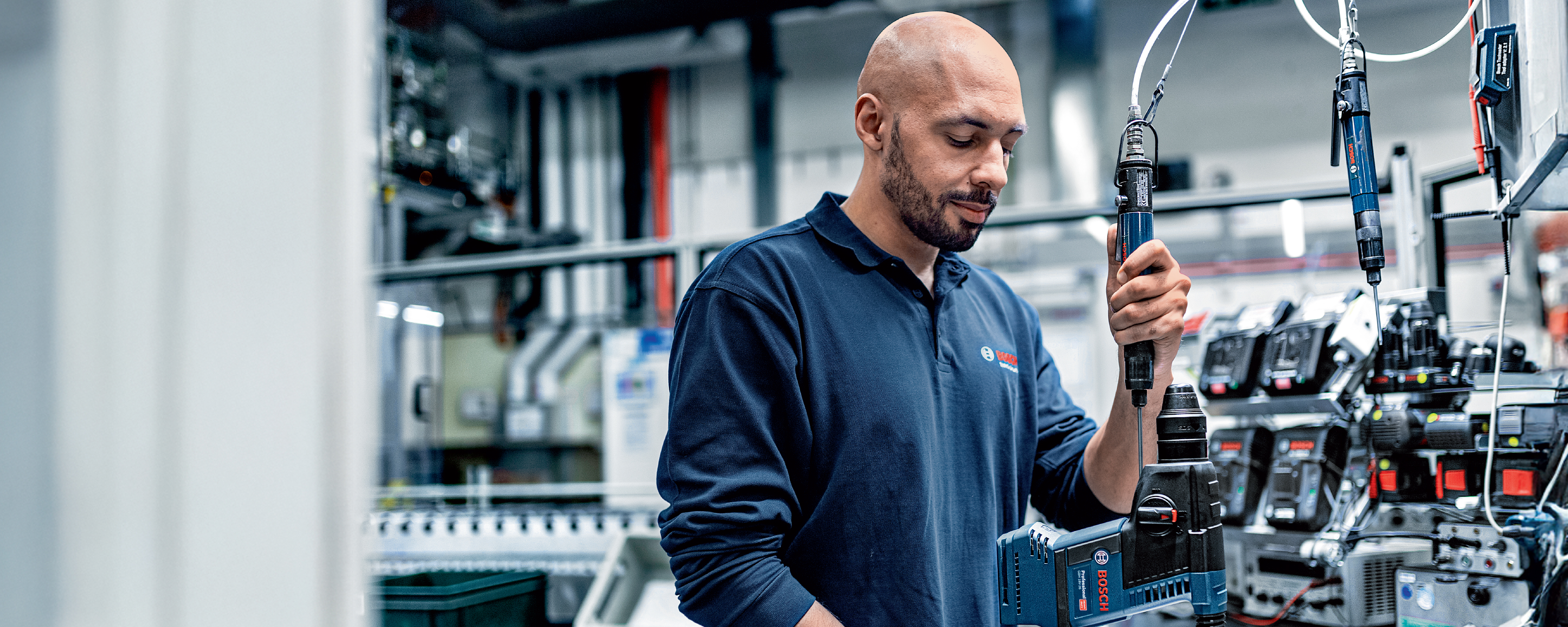 A man in work clothes holds a tool in a production hall, surrounded by machines.