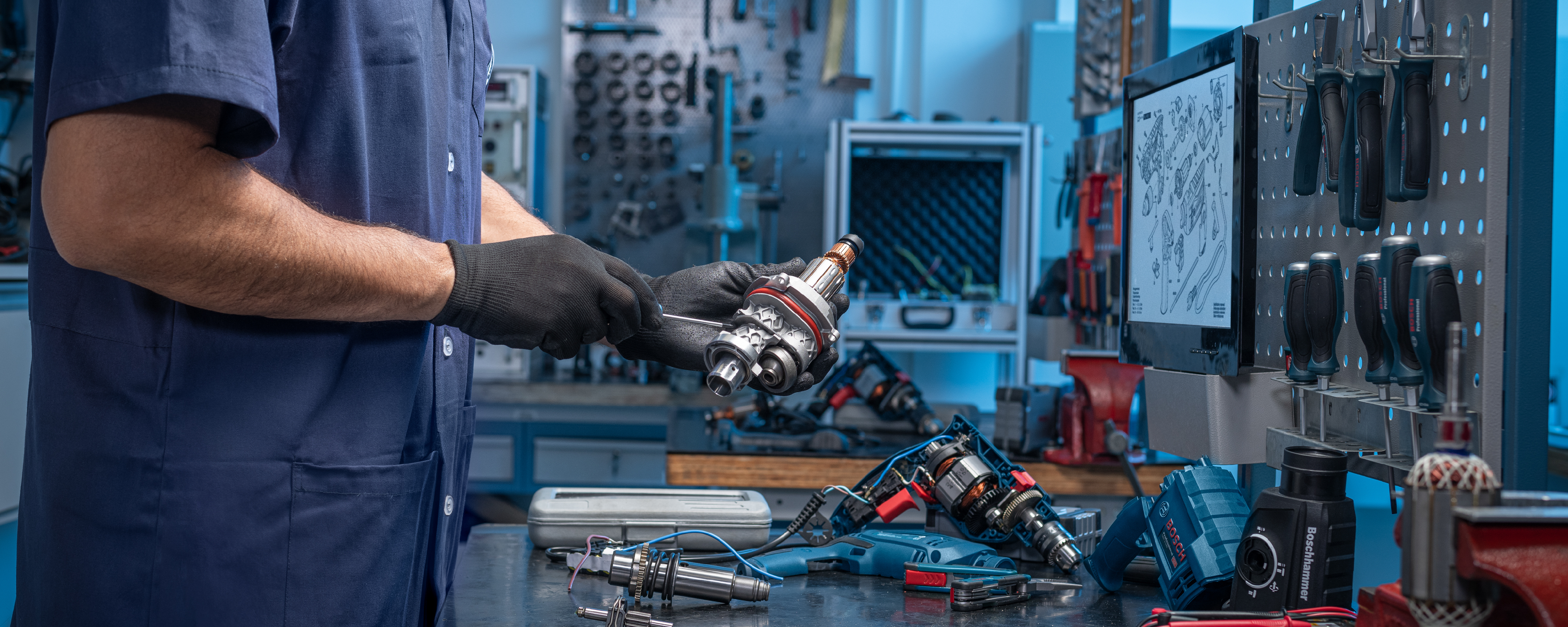 A mechanic in blue work clothes holds a metal part in his hand, surrounded by tools.
