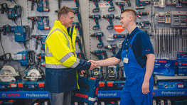 Dva muškarca se rukuju ispred Bosch polica sa alatima, jedan nosi žuto.