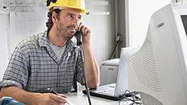 Мужчина в желтом шлеме говорит по телефону, сидя за компьютером.