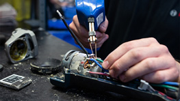 A person holds a blue soldering iron and works on an electrical device on a table. Various tools and components are visible, including cables and a multimeter. The environment is well-lit and conveys a craft atmosphere.