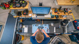 Stół warsztatowy z narzędziami, komputerem i osobą pracującą.