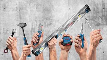 Des mains tiennent divers outils comme un marteau, un tournevis et un niveau à bulle devant un mur en béton.