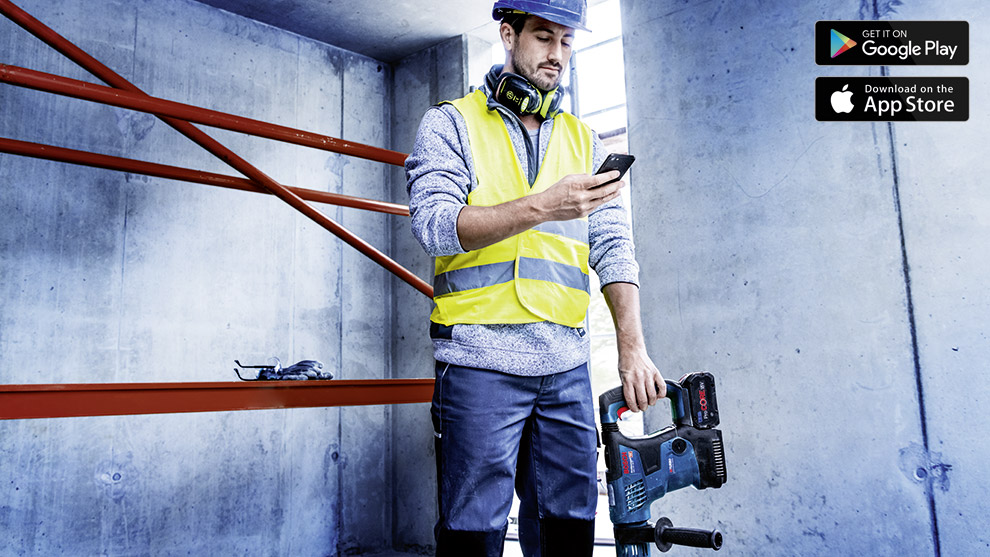 Čovek u reflektujućem prsluku i bušilici stoji u građevinskom okruženju.