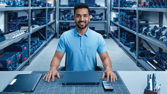 A smiling man in a blue polo shirt sits at a table in a tool warehouse, surrounded by shelves of Bosch Professional tools. On the table are two black folders and a smartphone.