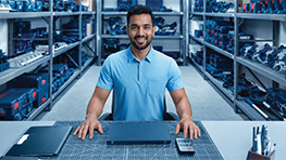 A smiling man in a light blue polo shirt sits at a work table in a workshop. In the background, there are shelves with Bosch Professional tools in various colors, mainly blue. The table is decorated with a gray work sheet and a black folder. The atmosphere is friendly and professional.