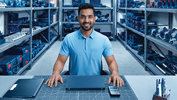 A smiling man in a light blue polo shirt sits at a table in a tool warehouse. In the background, there are shelves with Bosch Professional tools in various colors and shapes. On the table, there is a laptop, a tablet, and writing utensils neatly arranged. The atmosphere feels friendly and inviting. The floor is light, and the walls are neutral.